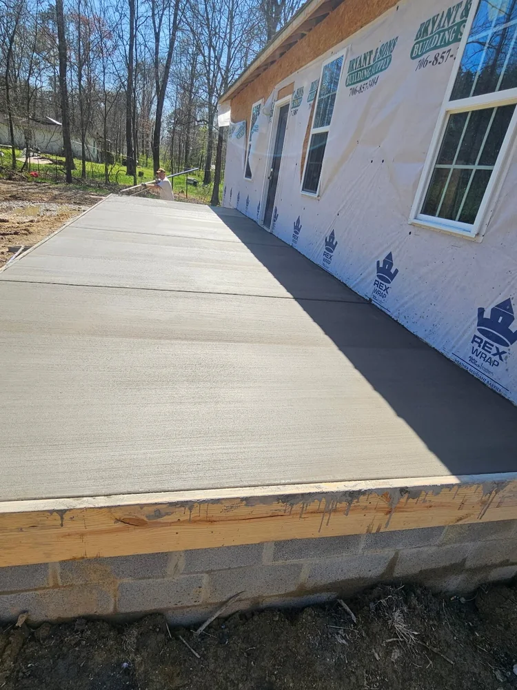 A broom-finished concrete walk poured the length of a house still in its building wrap, sitting on a gray block foundation
