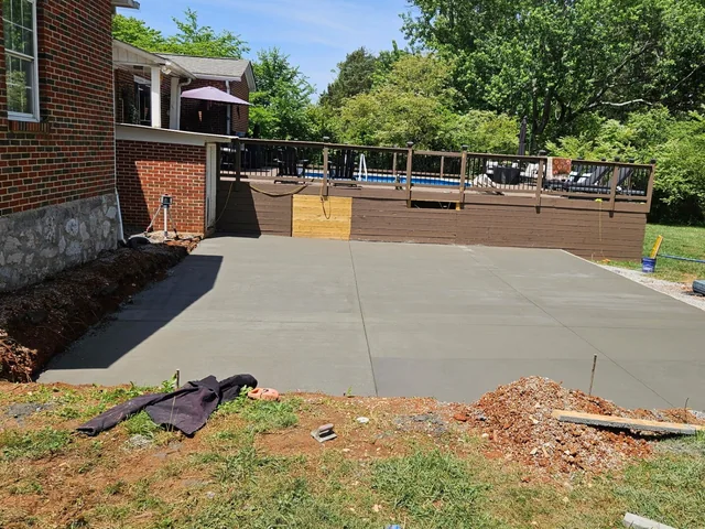 A freshly finished gray concrete pad in a back yard beside a brick house, with a raised wooden deck and an above-ground pool behind it