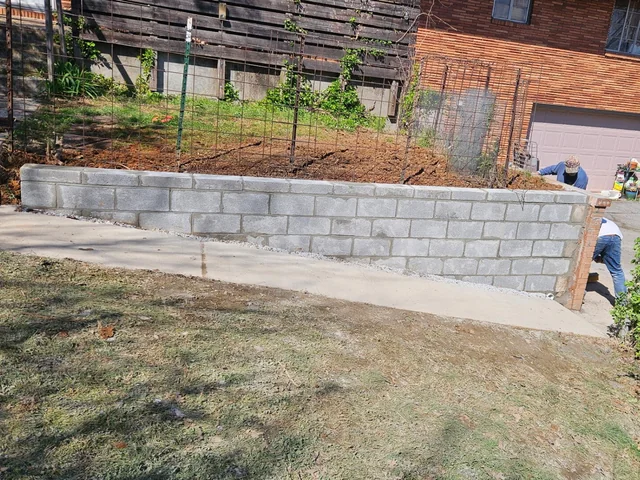 A long gray block retaining wall running across a yard below a dark timber building, with a poured concrete strip at its foot and a red brick house at the far end