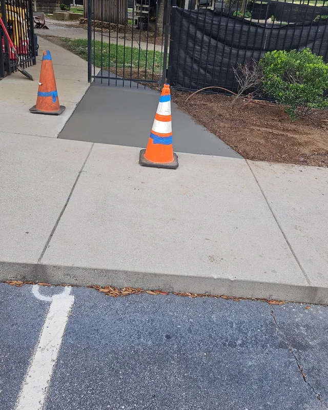 A new concrete sidewalk panel poured flush into an existing walk beside an iron gate, with traffic cones standing on it and a parking bay beyond the curb