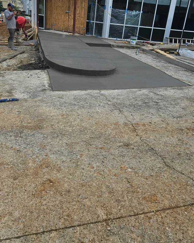 A fresh concrete entrance apron with a curved ramped edge laid at the doors of a glass-fronted building, a worker screeding at the far end
