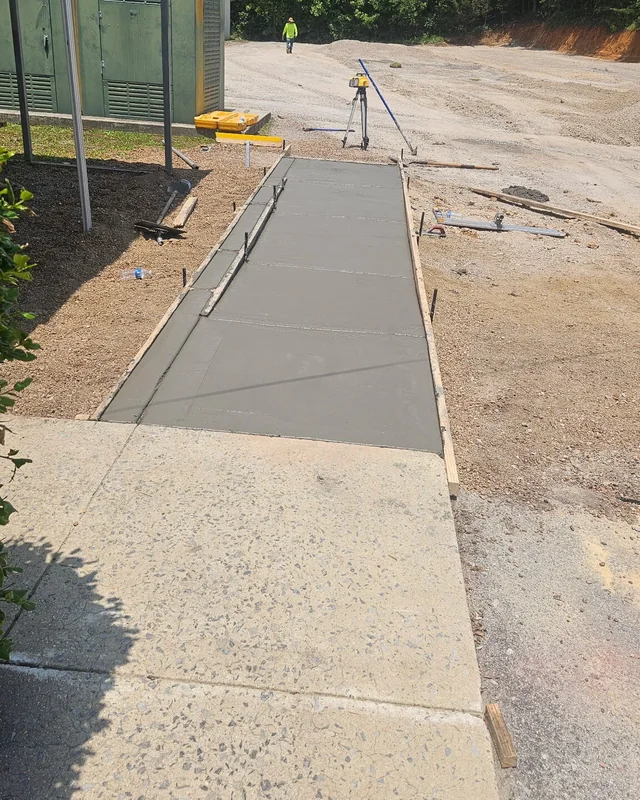 A long run of new sidewalk poured between timber forms beside a gravel lot, with a surveying level set up on it and a worker in hi-vis at the far end