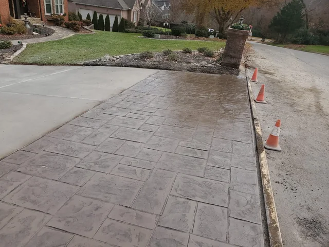 A wide stamped concrete driveway in a large ashlar pattern, wet and dark, running past a brick pier with traffic cones still set out along its edge