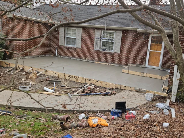 A freshly poured gray concrete patio behind a red brick ranch house with timber forms still pinned around it and offcuts and tools scattered across the bare ground