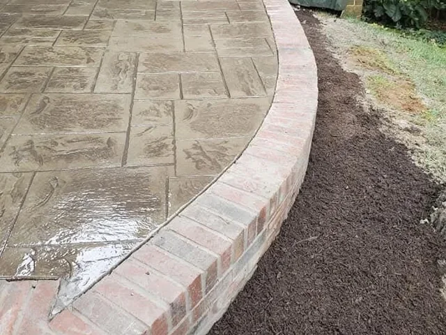 A wet stamped concrete walk curving away beside a house, with a single course of red brick laid flat along its outer edge and fresh mulch beyond it