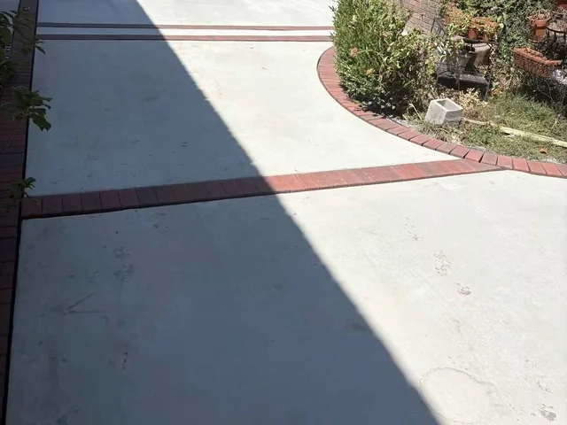 A back garden patio of pale gray concrete with a red brick band laid around its edge, curving away past a blue canopy and a brick wall at the top of the slope