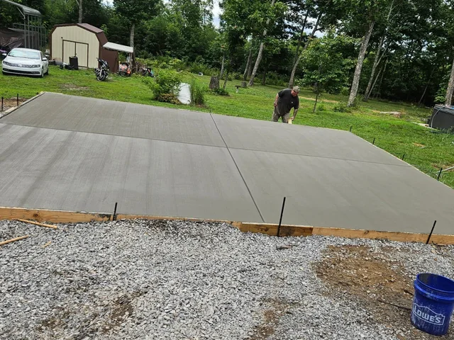 A large rectangular concrete slab finished smooth in an open back yard, timber forms still pinned around it and a gravel base showing in the foreground