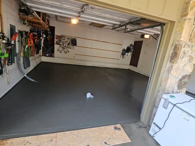A new concrete floor finished inside a garage with tools hung along the back wall, a stone-faced column at the opening and a strip of lawn outside