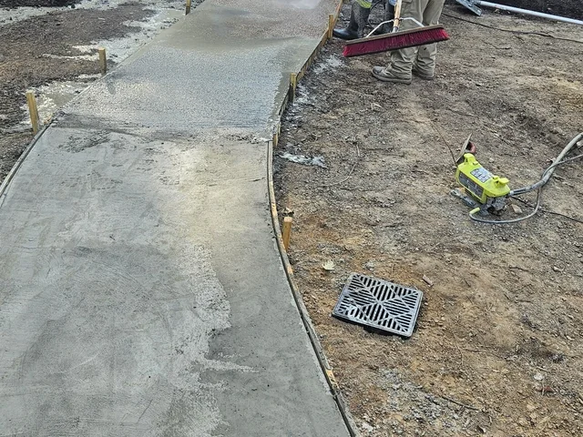A fresh concrete walk curving away across a site, a worker brooming the far end of it, with a drain grate and a power tool lying on the ground beside it