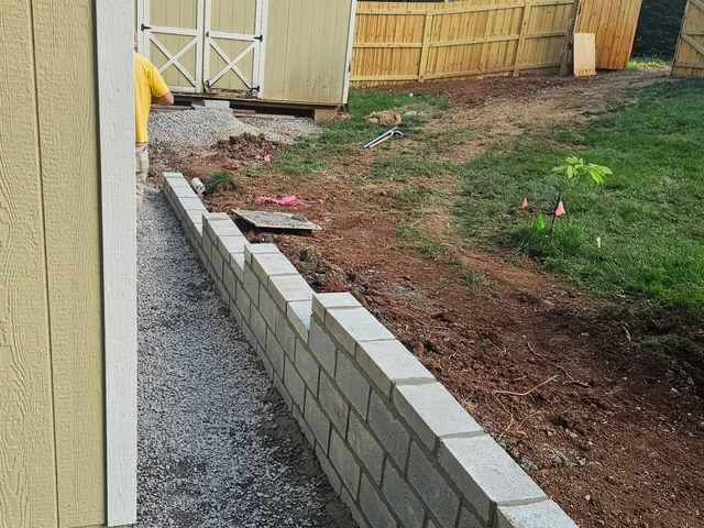 A gray block retaining wall running along the side of a shed, with gravel filled in behind it and raw red clay in the foreground
