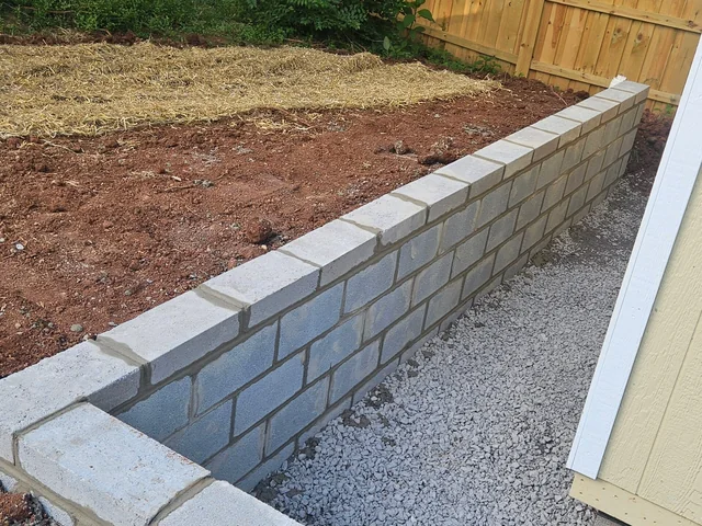 A gray block retaining wall built along a timber fence line with gravel laid against its base and fresh soil graded flat behind it
