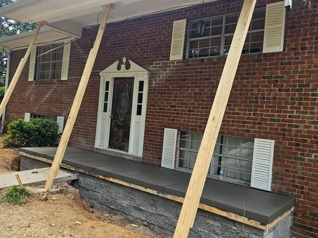 A new concrete porch along the front of a red brick house with timber props braced against it and the ground in front still open