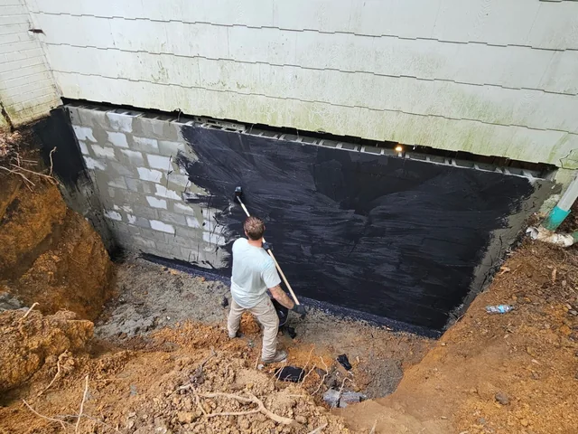 The outside face of a block foundation wall coated black with waterproofing down to the footing, with a worker standing in the open trench in front of it