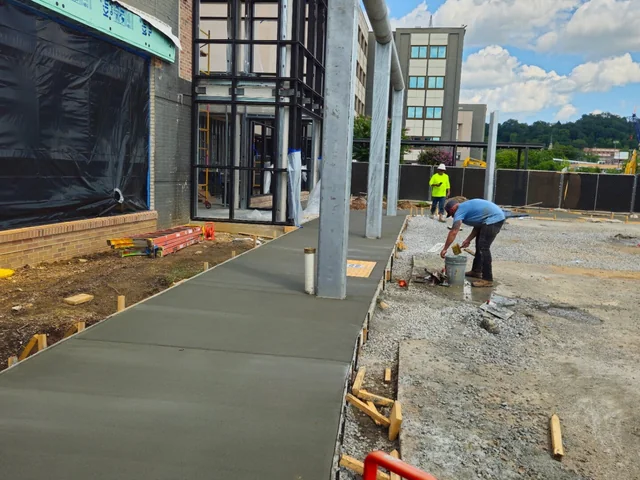 A new concrete sidewalk poured along the front of a glass-fronted building still under construction, with a worker finishing it at the far end