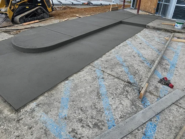 A new concrete ramp with a flared edge poured against an existing parking surface at the end of a building, with a machine working behind it