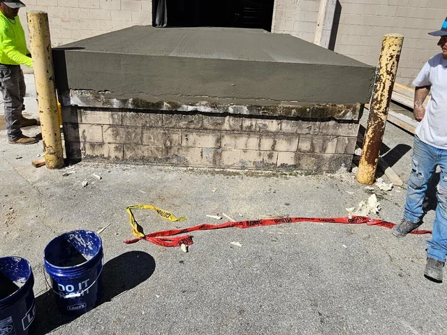 A loading dock whose stained and crumbling block base has been capped with a new concrete deck, with a worker standing behind it and scrap timber underneath