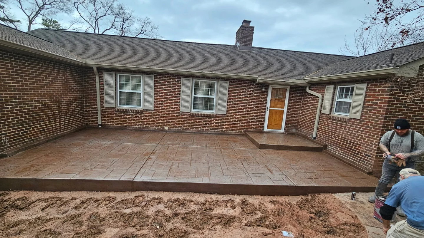 A stamped and colored concrete patio finished along the back of a red brick ranch house, with a raised step up to the back door and bare graded soil in front of it