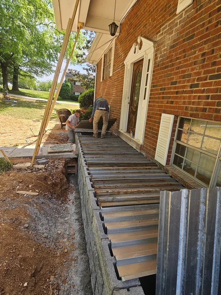 Timber forms set out and braced for a porch slab against the wall of a brick house, with the ground dug out in front of them