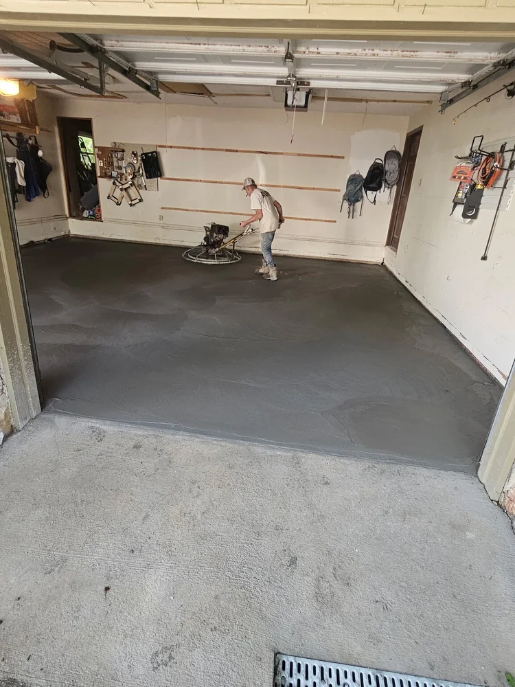 A worker power-floating a fresh concrete garage floor inside an open garage, with tools hung along the wall behind him
