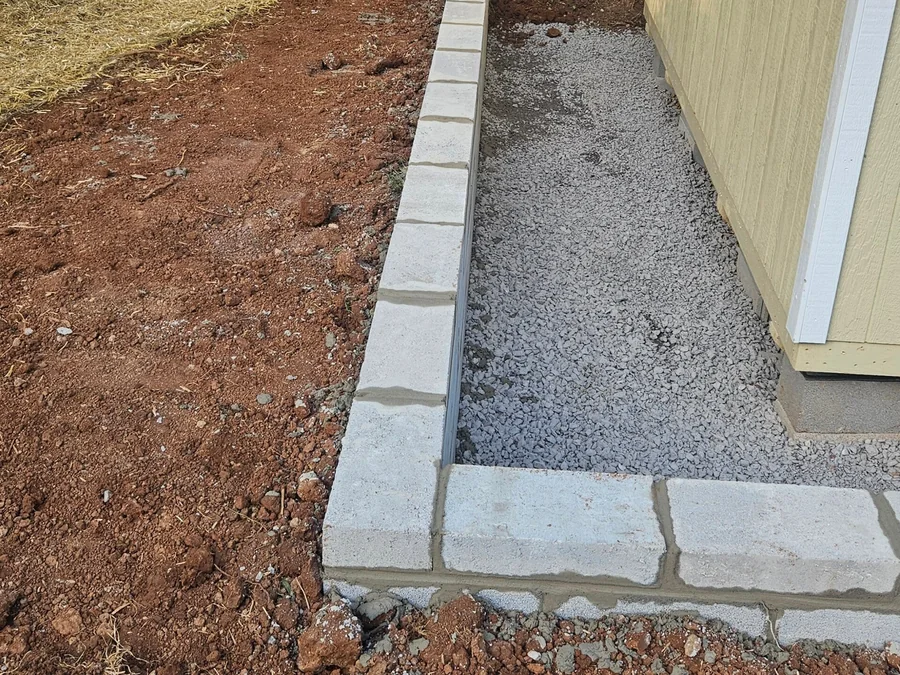 A gray block retaining wall returning around the corner of a yellow building with gravel filled in behind it