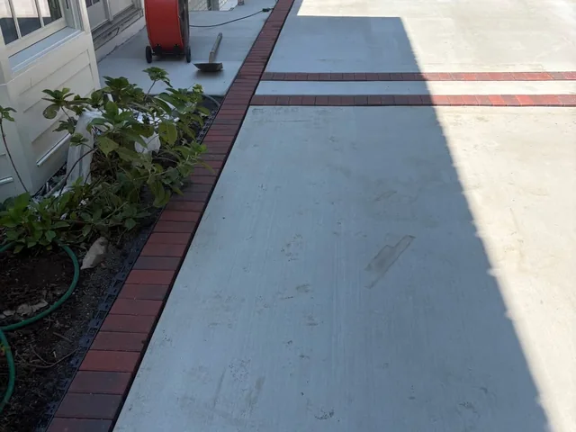 A gray concrete walk running along the side of a house with a red brick band down its outer edge and a second brick band laid straight across it, a hose coiled in the planting bed and a red machine standing at the far end