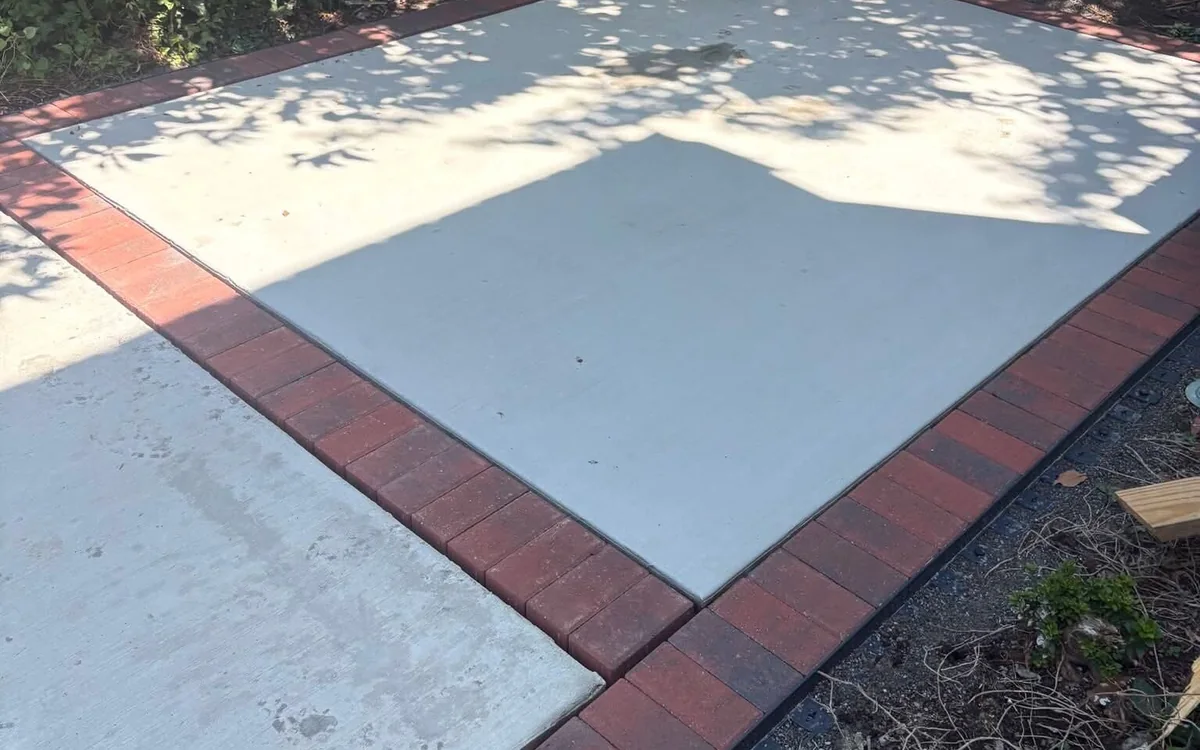 A square pad of pale concrete framed on every side by a single course of red brick laid flat, set into bare ground at the back of a garden