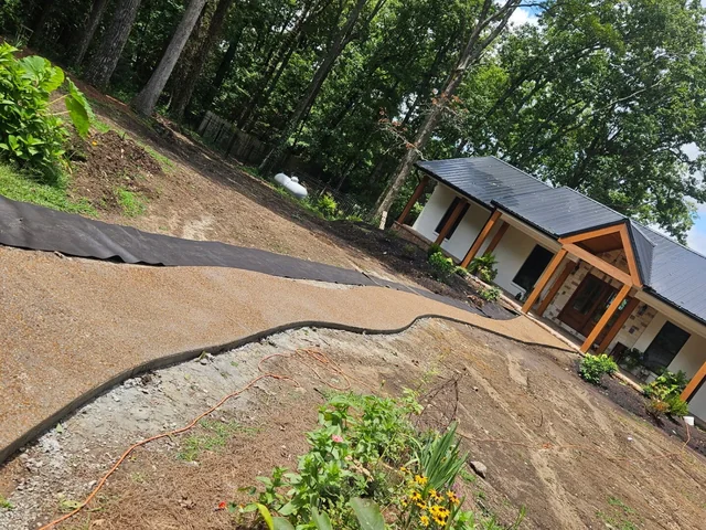 A broad tan concrete drive sweeping in a long curve in front of a white house with timber porch posts and a dark metal roof, black landscape fabric pinned down the bank above it and planting going in along the near edge