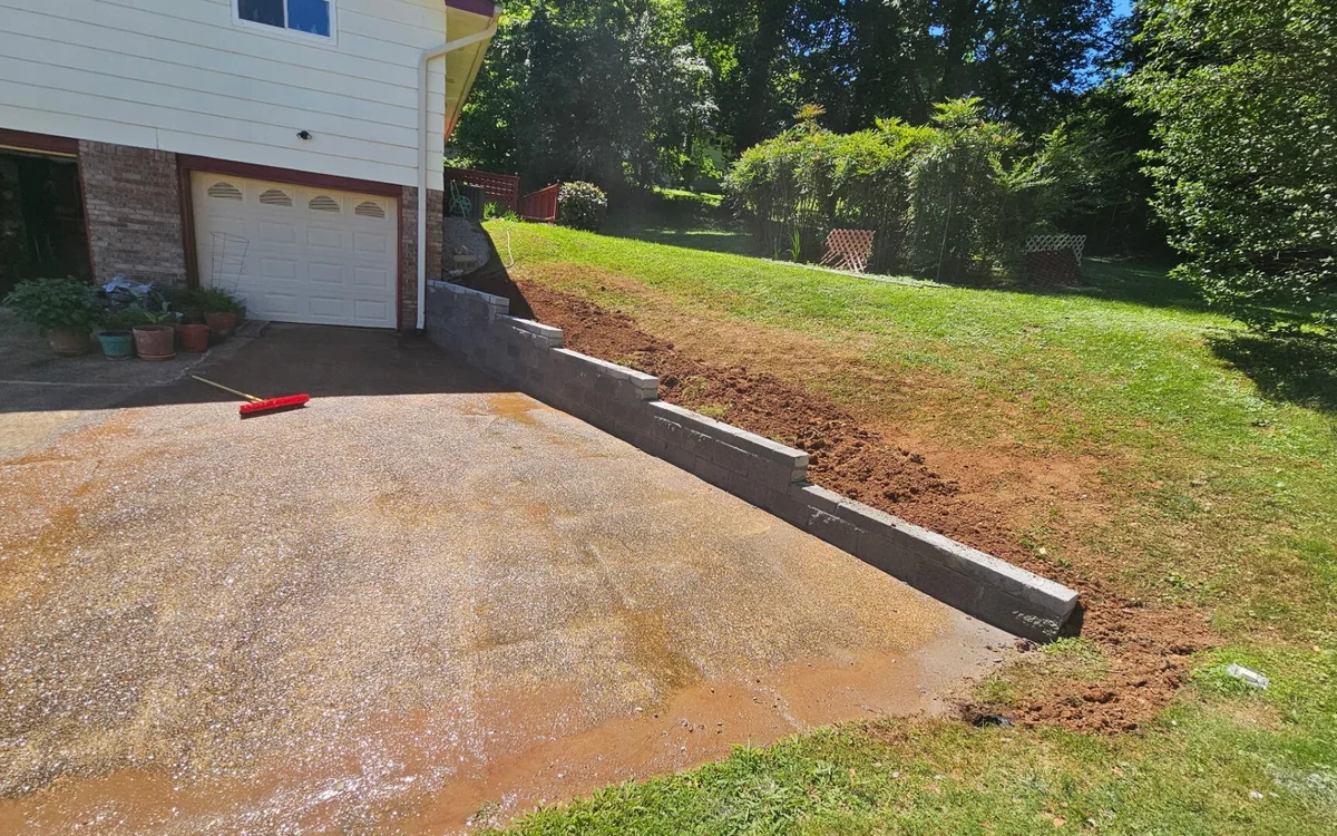 A wide driveway in wet reddish-tan concrete running up a slope to a white two-car garage door, with a low poured concrete retaining wall along its right-hand edge holding back the lawn above it and a red push broom lying on the surface