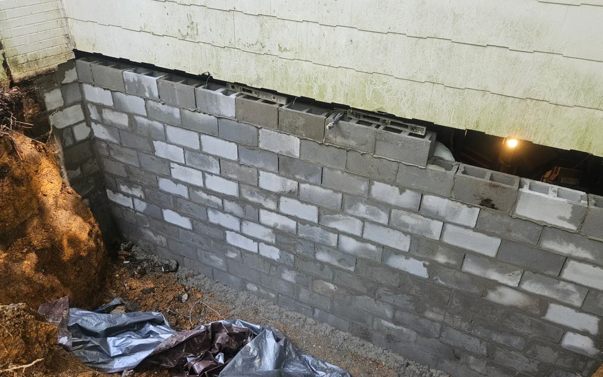 A block foundation wall exposed by excavation under a white house, a course of new gray block set into the top of it where the old block was taken out, with sheeting laid over the spoil
