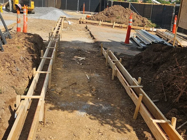 Two long runs of timber footing form set out in a trench on a city site, braced and staked along their length, with traffic cones, a spoil heap and a tracked machine behind them