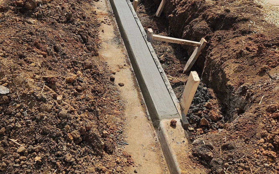 A poured concrete footing running away through a trench cut in red clay, timber kickers staked against its outside face to hold the form while it set