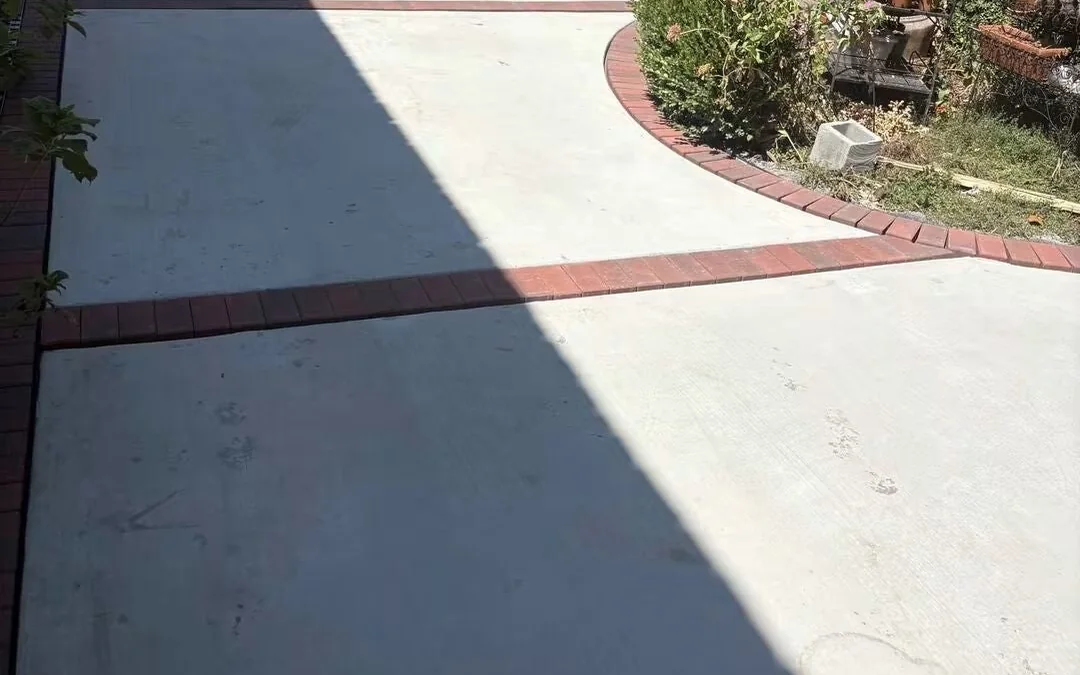 A back garden patio of pale gray concrete with a red brick band laid around its edge, curving away past a blue canopy and a brick wall at the top of the slope