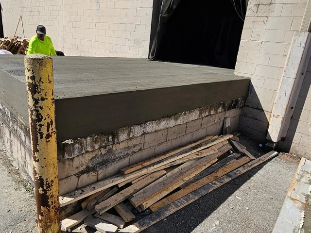 A loading dock whose stained and crumbling block base has been capped with a new concrete deck, a worker in a hi-vis shirt and cap standing behind it, a rusted yellow bollard in front and scrap timber stacked underneath
