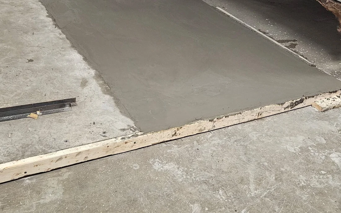A fresh pale slab poured hard up against an older, darker concrete floor, the joint between the two running straight across the frame, with a timber form and a length of steel angle still lying along the near edge