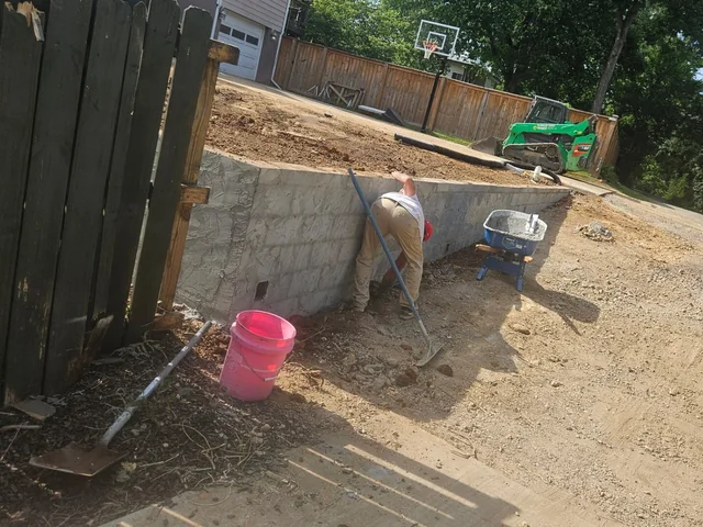 A worker digging at the foot of a part-built gray block wall against a dark timber fence, a wheelbarrow and a pink bucket beside him and a skid steer parked at the top of the slope
