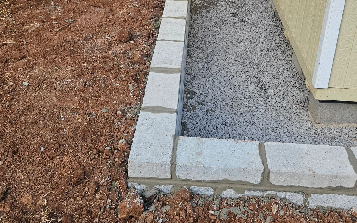 A low gray block retaining wall returning around the corner of a yellow-sided building, washed gravel filled in between the wall and the wall of the building, and raw red clay in the foreground
