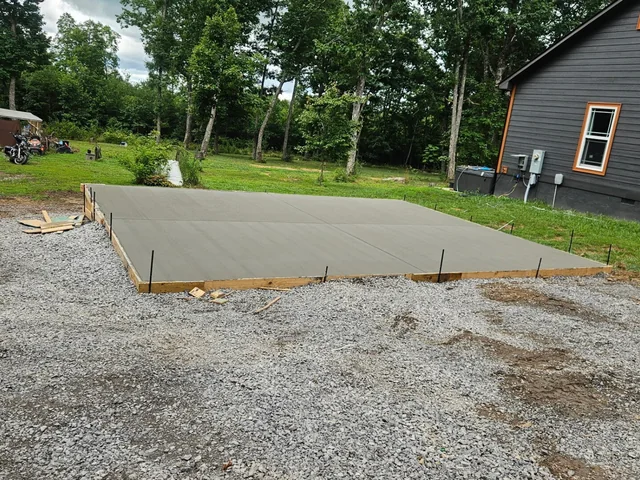 A slab poured level with its timber forms on a gravel base, the ground around it still open, with a gray-sided house and an electrical panel behind it