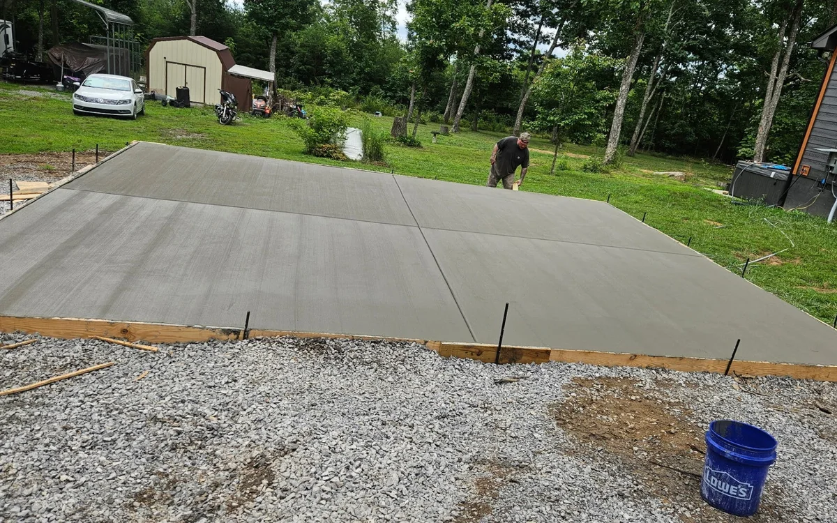 A large rectangular concrete slab finished smooth in an open back yard, timber forms still pinned around it and a gravel base showing in the foreground, with a shed and trees behind