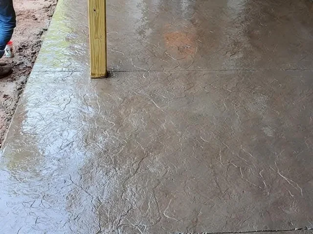 A wet stamped slab running away from a timber post, the surface mottled tan and gray where the color has not yet dried back, with raw earth and somebody's boot at the top edge of the frame