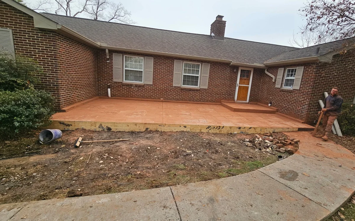 A newly stamped patio behind a red brick ranch house still carrying its orange color release, timber forms along the front edge and a curved concrete walk running past it