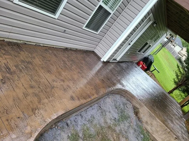 A stamped and colored concrete walk being washed down alongside a gray-sided house, the surface wet and dark, with a green pressure washer standing on the lawn beside it