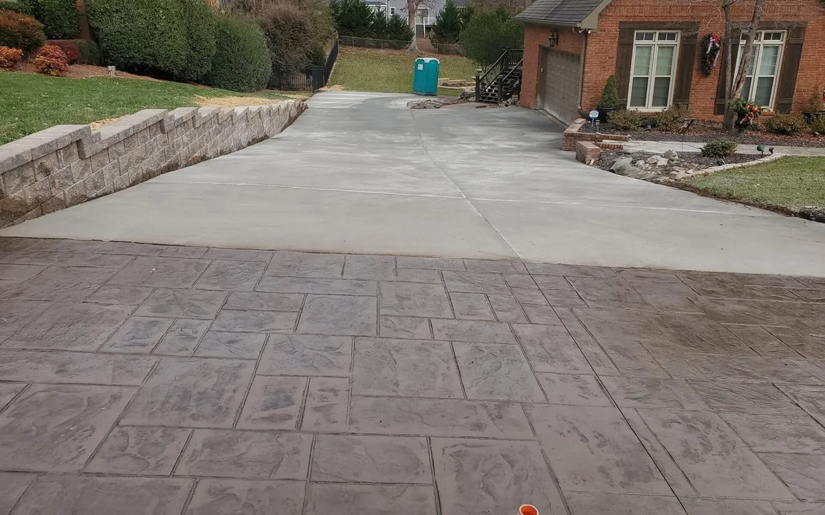 A stamped concrete driveway in a large ashlar pattern running up to a red brick house, the plain gray driveway it joins visible beyond it and a gray block wall down the left-hand side