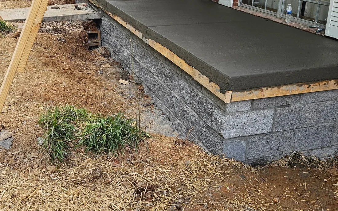 A new concrete porch slab finished flat on top of a gray block stem wall, timber forms still pinned along its edges, with straw spread over the bare ground in front and a clump of grass left standing beside it