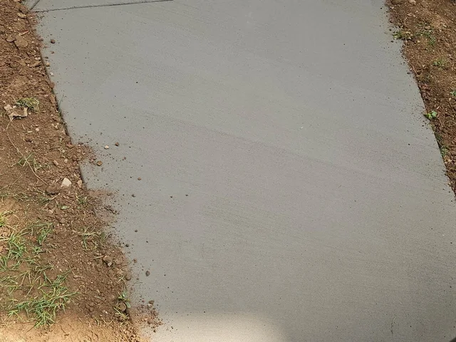 A plain gray concrete walk running between a lawn and a planted bank, jointed across its width, with a timber bench and a dark green building at the far end