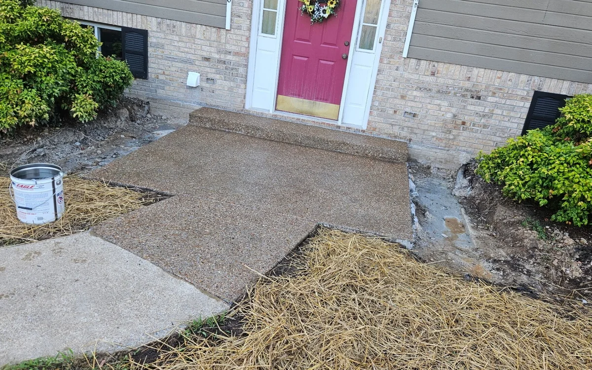 A concrete front walk and landing leading to a pink front door on a brick and gray-sided house, with fresh straw spread over the bare ground either side of it
